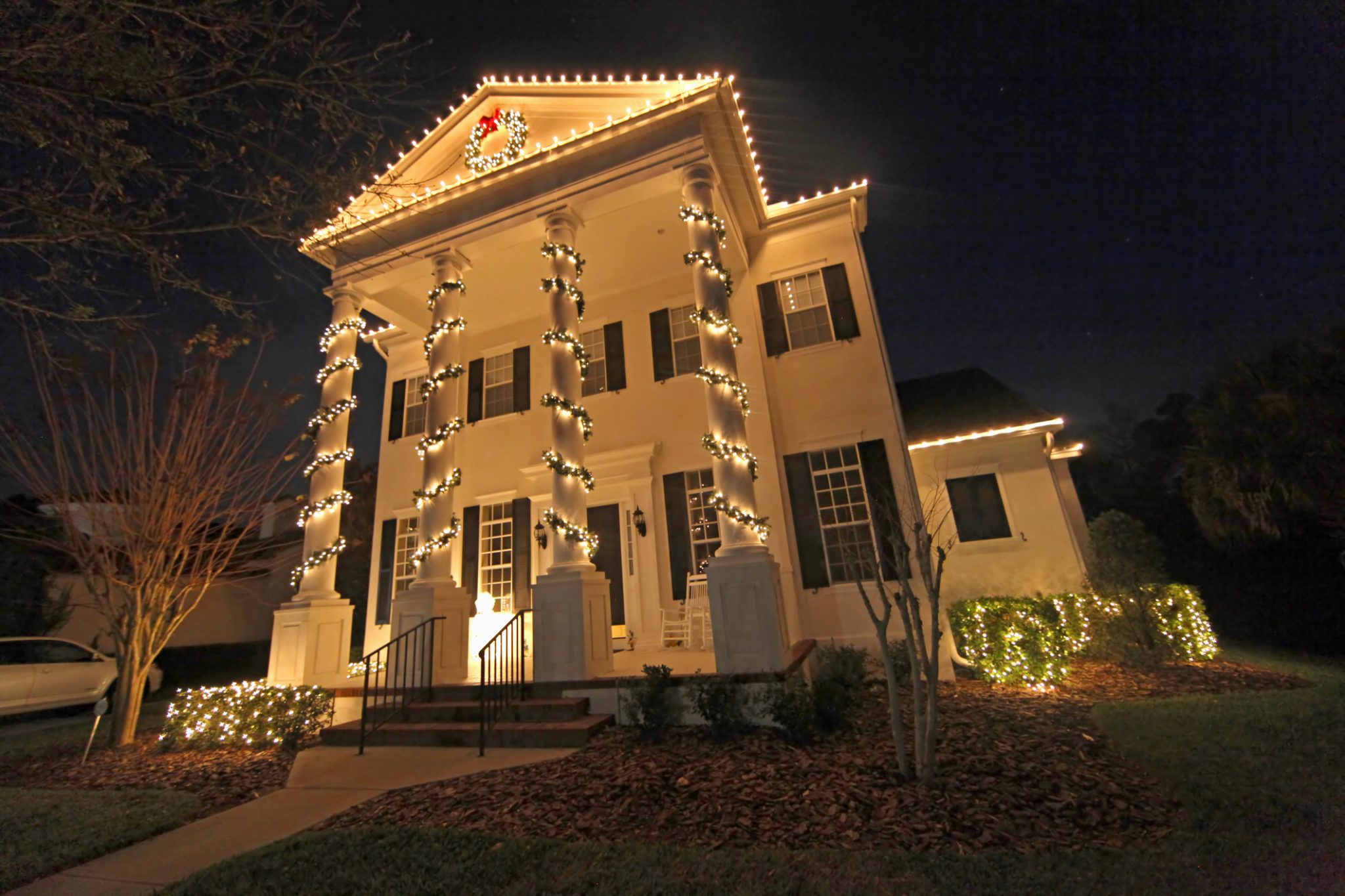 Large southern-style home with pillars wrapped in lights and roofline outlined in warm white holiday lights.