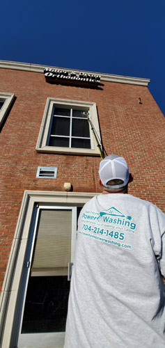 Technician from Clean Home Power Washing cleaning second-story window with professional equipment on a brick commercial building.
